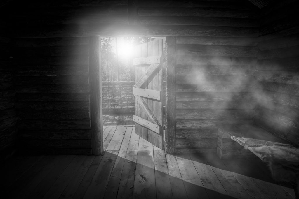 Black and white photo of an old log cabin in the dark with light coming in from an open door. 