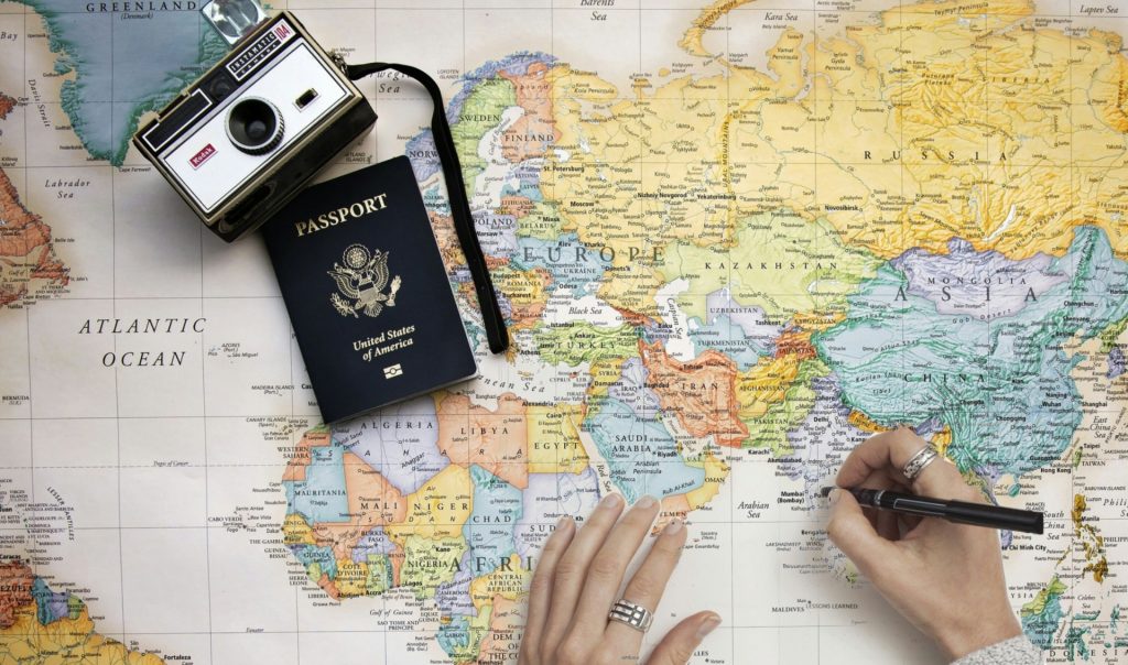 Image of a person's hands wearing rings and drawing to mark places on a map. There's a passport and a camera on the map and they're making a mark close to India. 