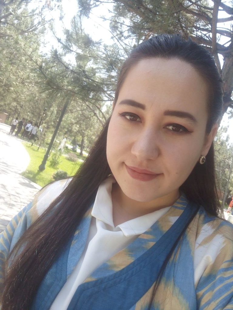 Central Asian young woman with long dark hair, earrings, and a blue, tan, and white coat over a white collared shirt. She's in a park with trees and a lawn and lamp. 