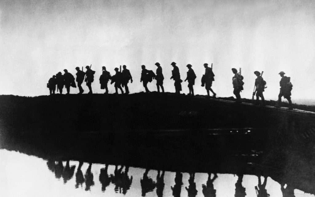 Black and white photo of a line of soldiers carrying packs and rifles marching past a body of water. 