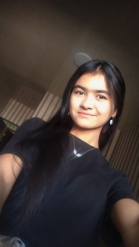 Selfie of a teen Central Asian girl with long dark hair, a black tee shirt, a silver necklace and earrings. 