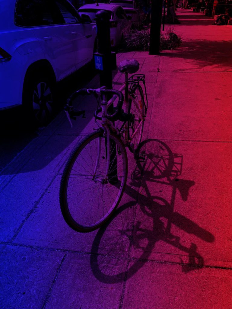 Bicycle near a parking meter on a sidewalk at night that's red and purple from lights. White SUV and other cars parked. 