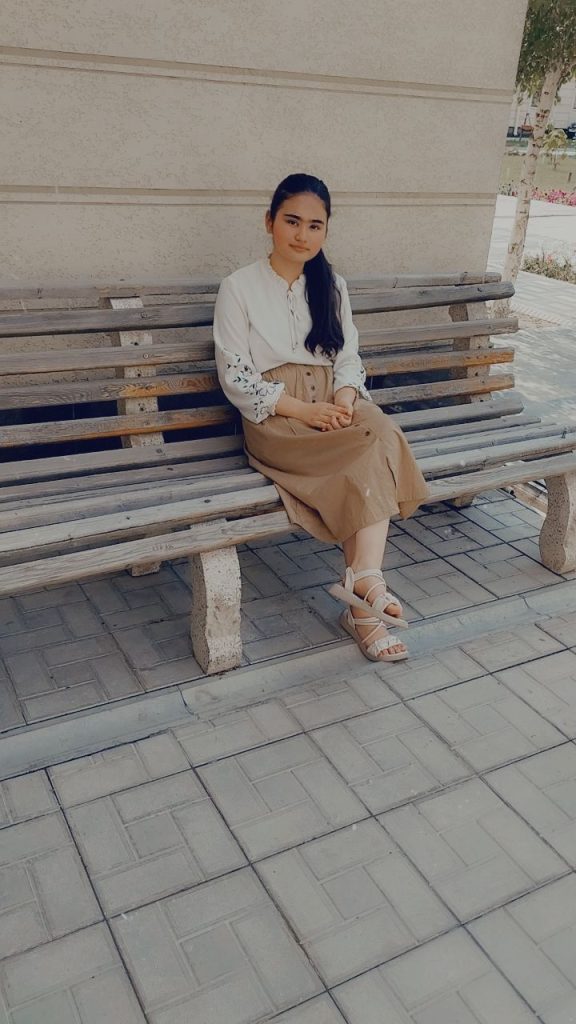 Young Central Asian woman in a cream colored blouse and tan skirt with long dark hair sits on a wooden bench outside a building. 