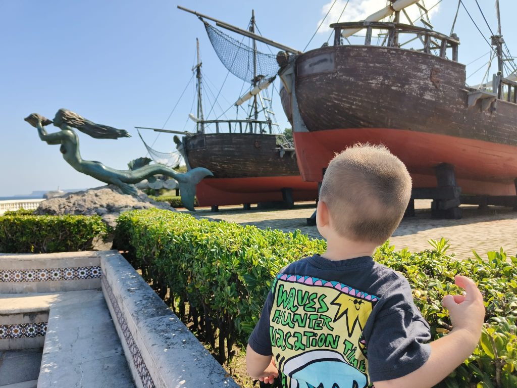 Small light skinned boy in a tee shirt facing a large wooden ship with a mermaid figure at the bow. Green bushes nearby. 