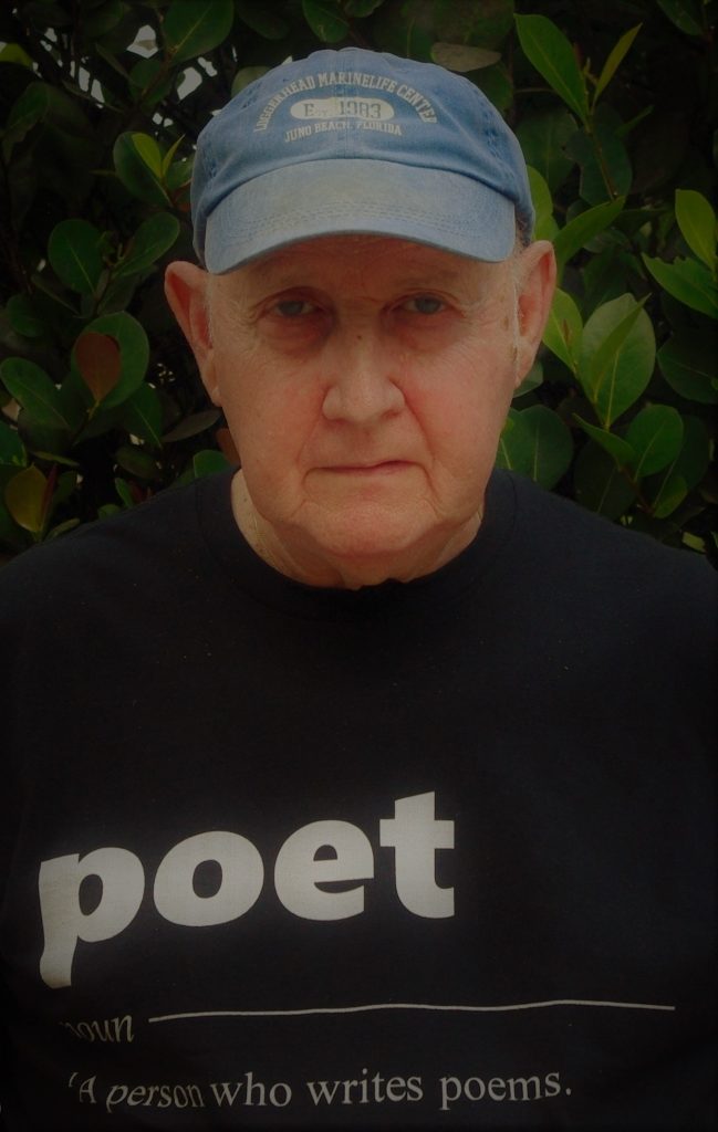 Older white man with a light blue baseball cap and a black tee shirt in front of a leafy bush. His shirt reads "poet, noun, a person who writes poems." 