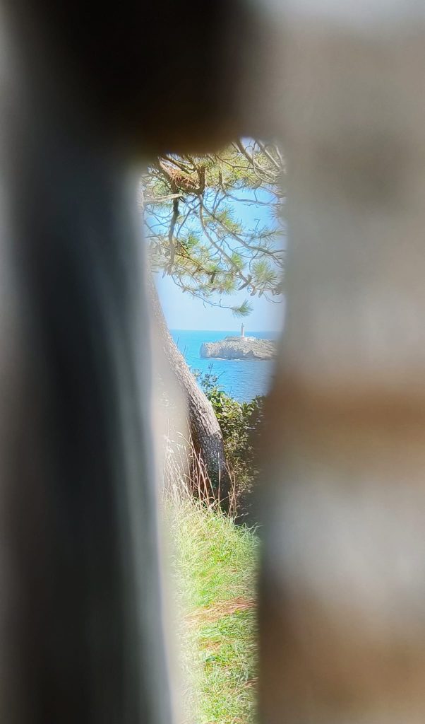 View of a lighthouse on a rocky outcropping near some pine trees and grass, through a small hole. 