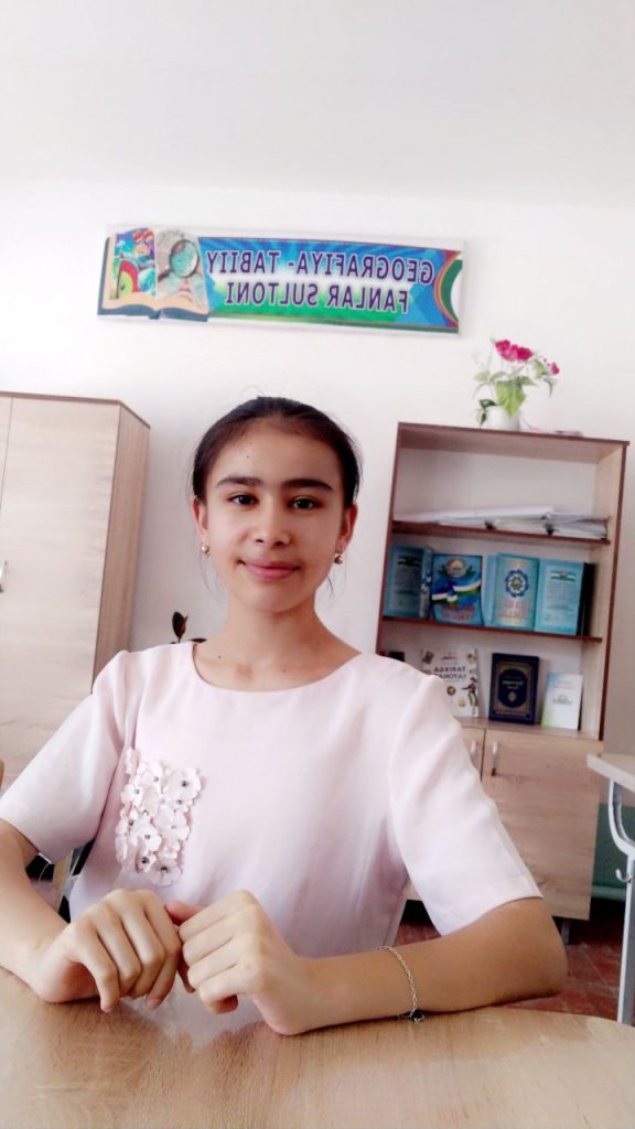 Central Asian teen girl with hair behind her head, small earrings, and a pink blouse with flowers on the right. She's seated at a desk with books on a shelf and flowers behind her. 