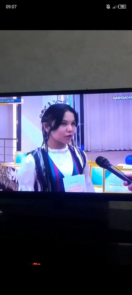 Teen Central Asian girl with an embroidered headdress, long dark hair, a colored vest and white blouse speaks into a microphone at an awards ceremony. 