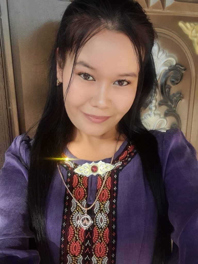 Teen Uzbek girl with long dark hair in a purple blouse with red and white embroidery, seated in a wooden chair.