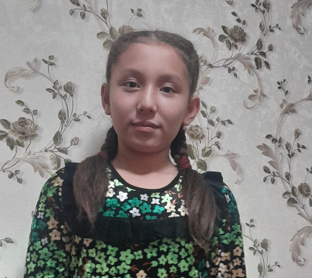 Central Asian teen girl with two braids of brown hair, a green and tan floral blouse, against floral wallpaper.