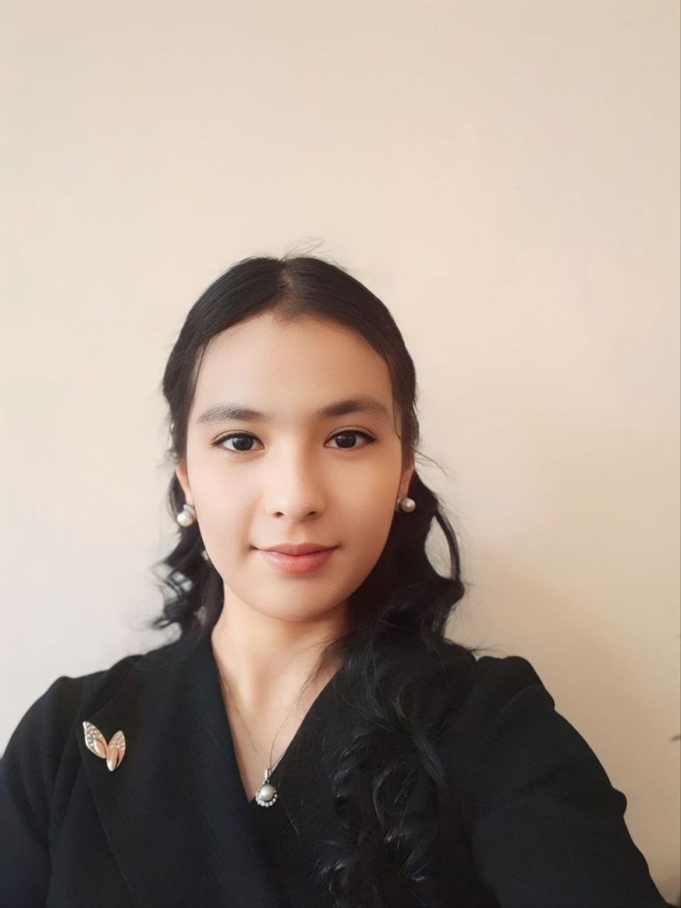 Young Central Asian woman with curly dark hair and a small necklace and earrings and a black top.