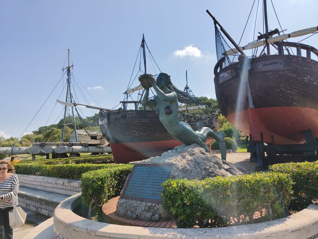 Two wooden sailing ships and a mermaid statue in green copper blowing on a horn. She's near a plaque and some green bushes. 