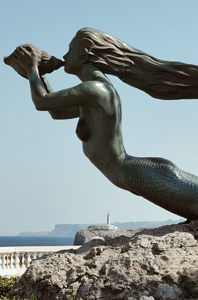 Closeup of the mermaid with long flowing hair blowing on a seashell. Lighthouse on a rock in the distance.