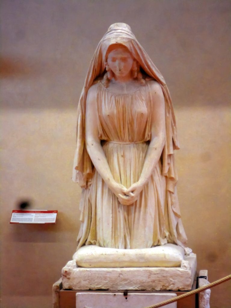 Statue of a veiled woman in a dress with curly hair kneeling over a grave. 