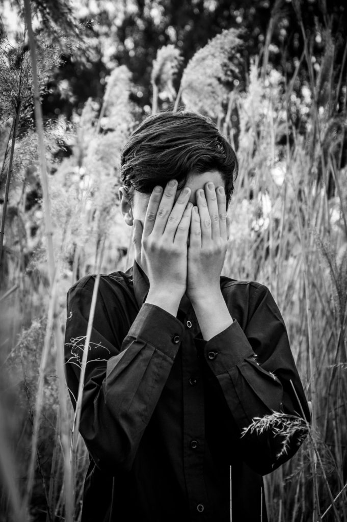 Young white guy with his hands over his face. Black and white image where he's standing in a field of tall grass and reeds. 