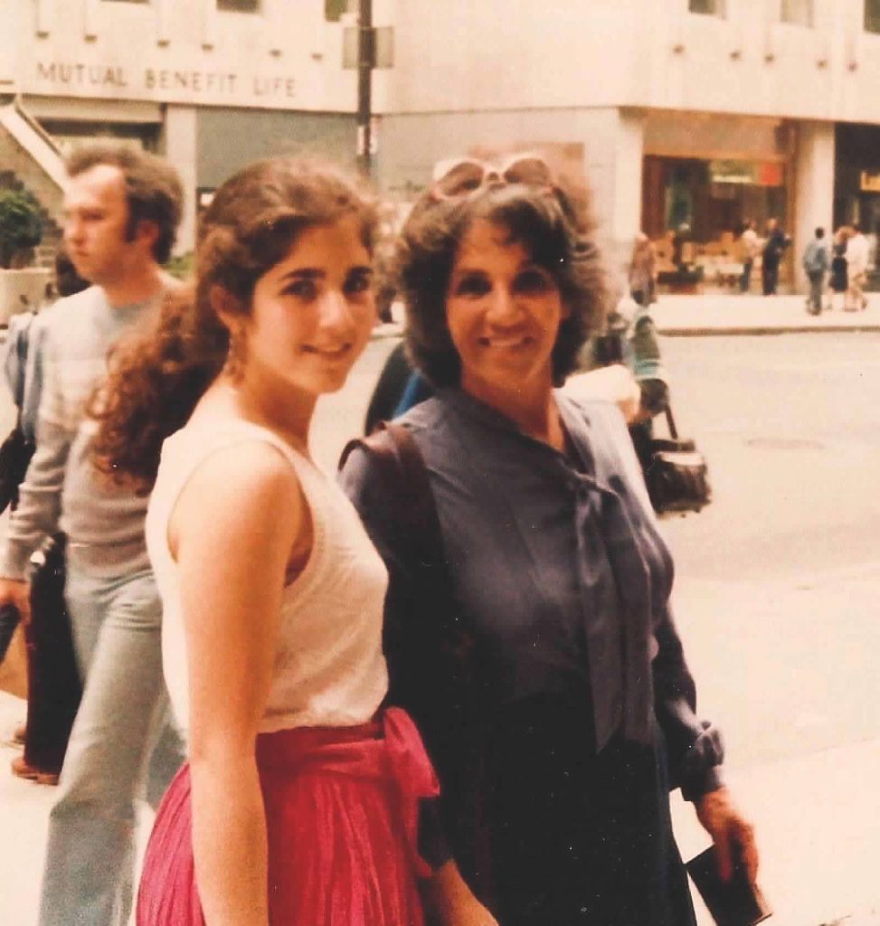 Teen light-skinned girl with curly dark hair and a white tank top and pink skirt stands outside in a street next to a young middle aged woman with a gray tied blouse, brown hair, and sunglasses. 