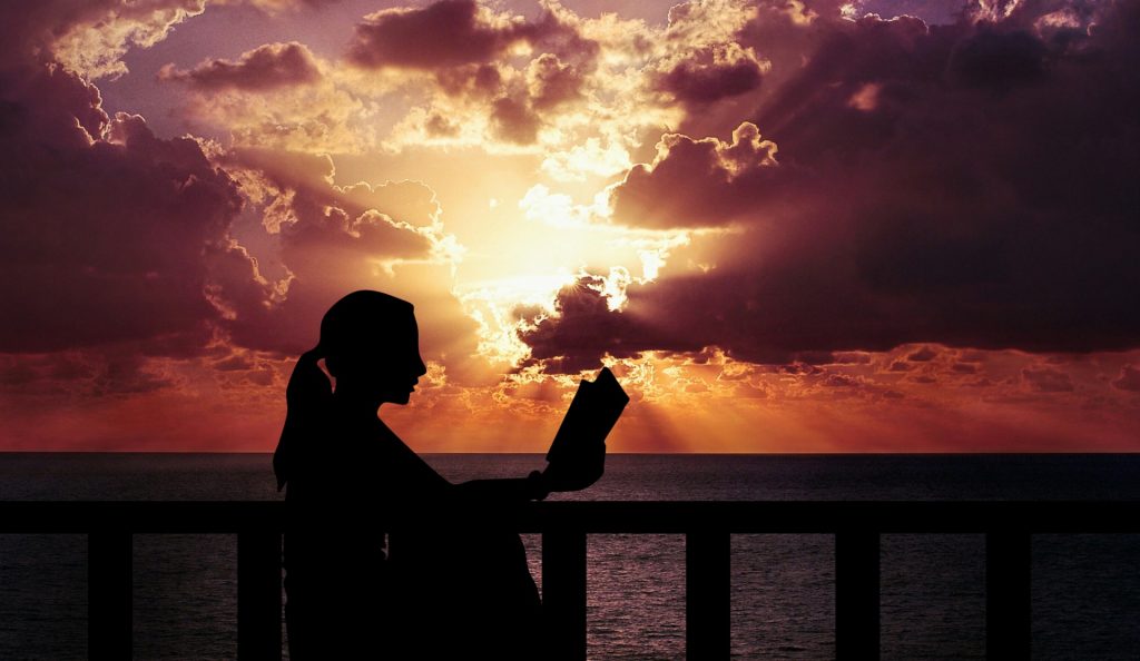 Woman with a ponytail of indeterminate race (silhouetted) reading a book by the ocean on a pier. Sun shines through clouds at sunset or sunrise. 