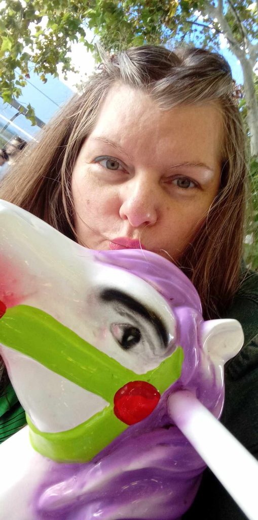 White woman with hazel eyes and light brown hair seated on a white, purple and green carousel horse. 