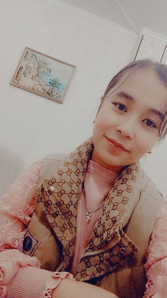 Central Asian teen girl in a patterned scarf and pink blouse and earrings. A small painting is in the background and she's leaning to the right. 