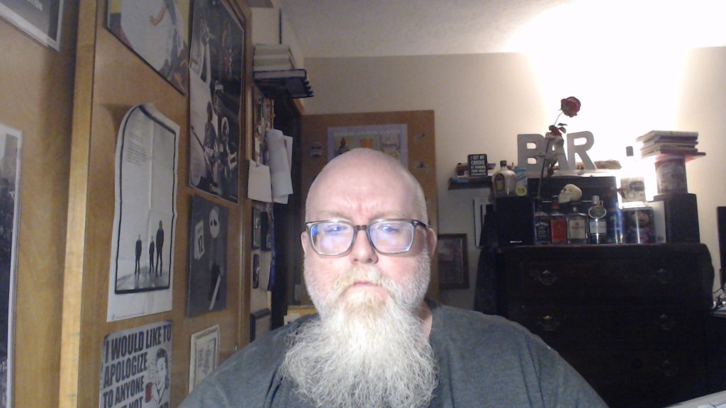 White middle aged man with a shaved head, long white beard and reading glasses. He's in a room with posters on the wall and a dresser with liquor bottles behind him. 