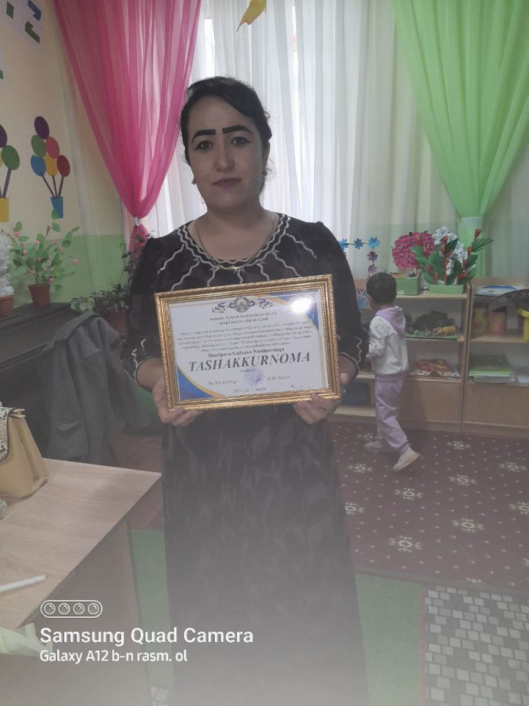 Central Asian woman with dark hair up in a bun, dark eyebrows, a dark fluffy blouse and skirt, holding a certificate. A child plays in the room behind her, toys and flowers nearby and green and pink and white curtains open. Balloons are painted on the wall, it looks like a child's playroom. 