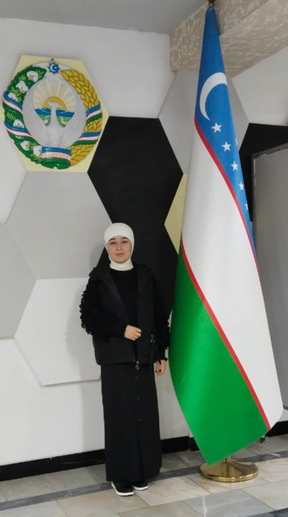 Young Central Asian woman with a black coat and white headscarf standing next to the Uzbek flag and a medallion with sheaves of wheat and white flowers. 