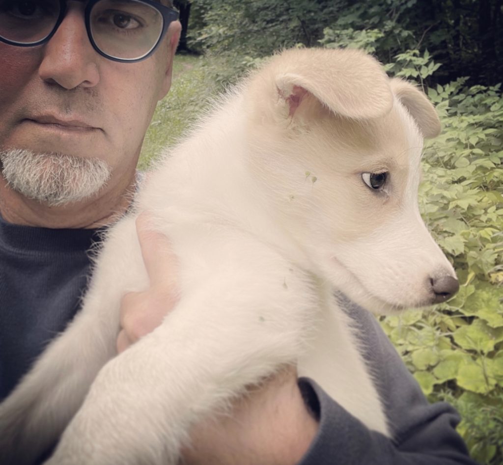 Middle aged white man with reading glasses and a tiny beard outside holding a furry white dog. Leafy bushes behind him. 