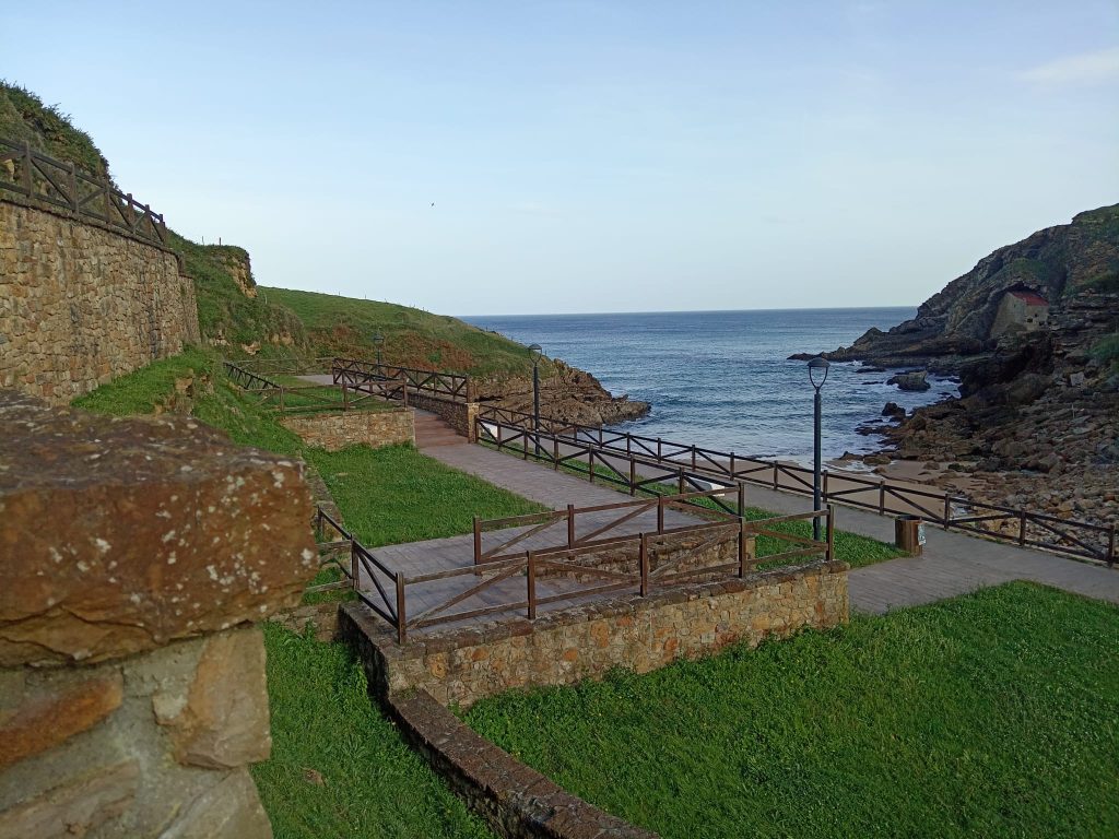 Stone walkway near some grass by the beach. Sand and rocky outcropping out at sea, stone wall on land. 