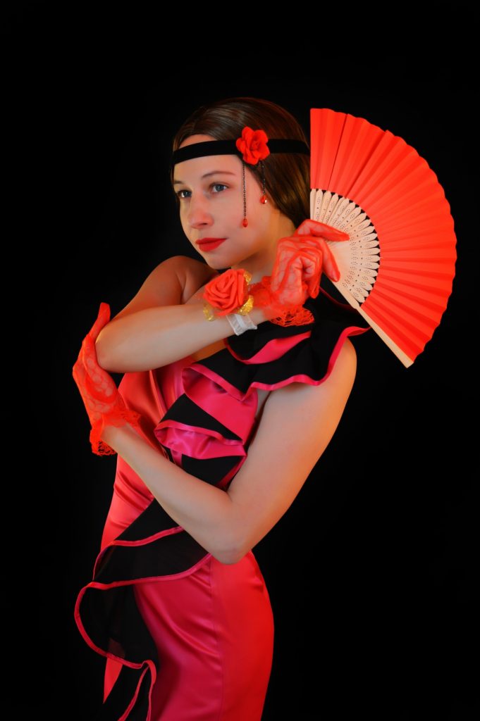 Profile from the side of a young light skinned woman with dark hair in a fluffy red and black tango dress and roses on her wrist and red gloves. 