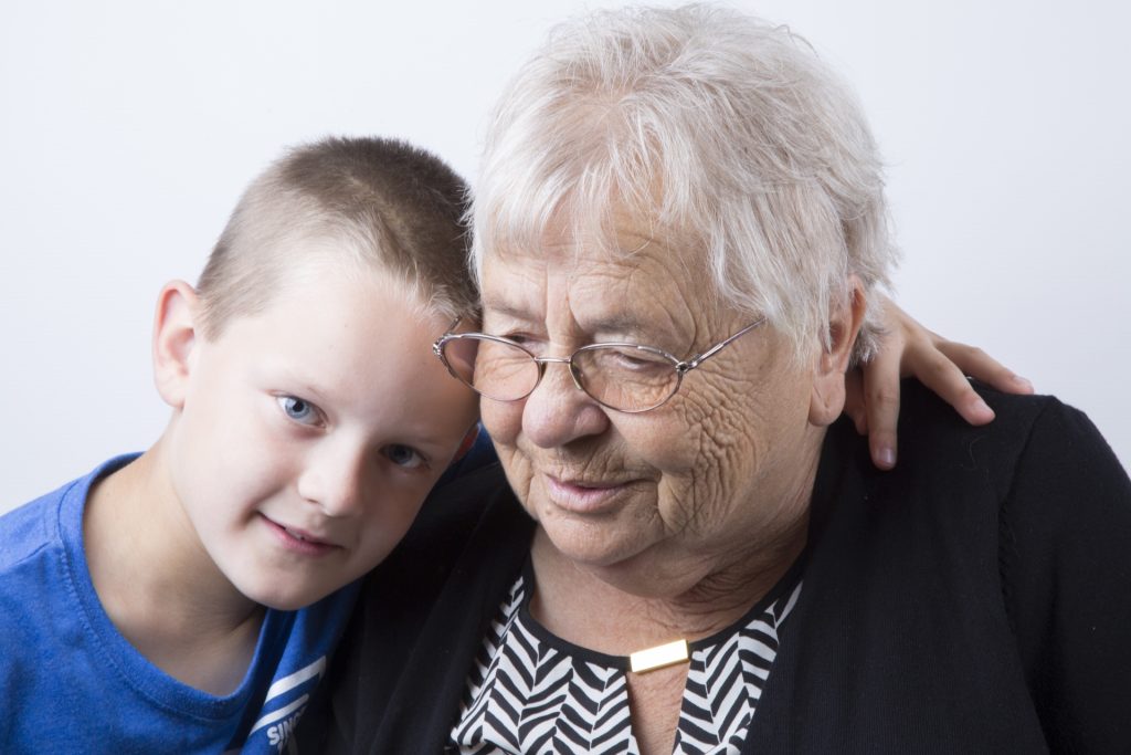 Older light skinned woman with reading glasses and a black sweater embraces a young blue eyed boy with short hair and a blue tee shirt. 