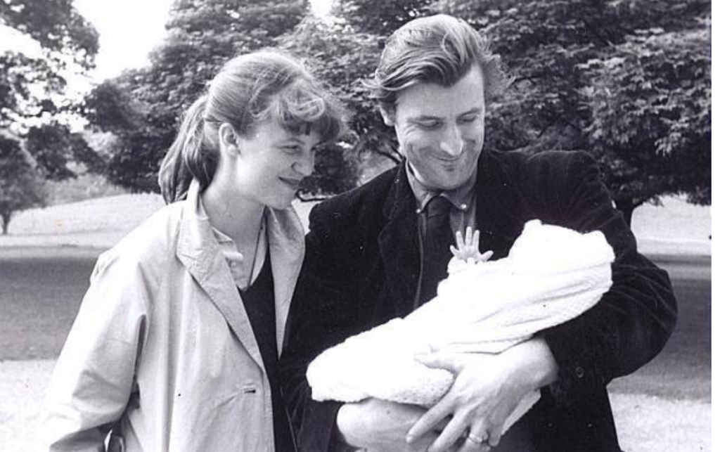 Young couple in jackets out under a tree holding a baby, who's raising its hand. 