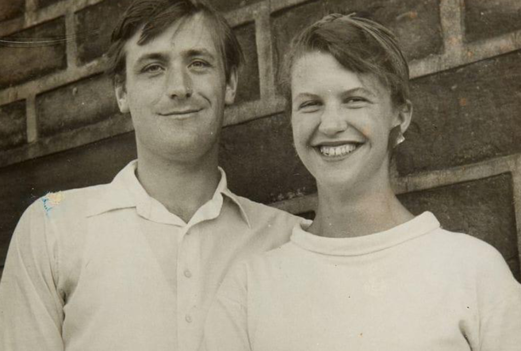 Young white couple with smiles and short brown hair pose in front of a brick wall. Both are in white collared shirts.