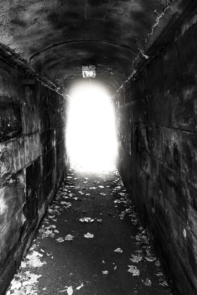 Black and white image of an old musty concrete tunnel with a light at the end.