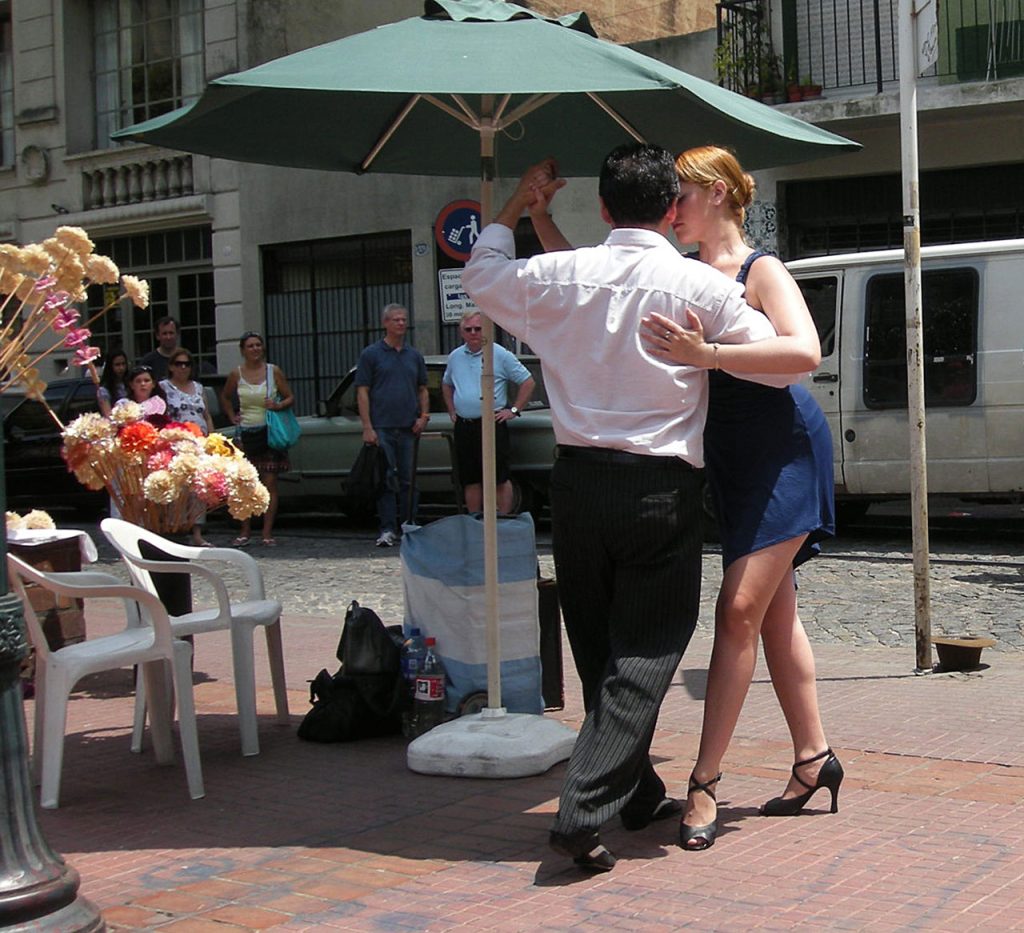 Realistic photo of a man and a woman dancing tango under a green umbrella on the sidewalk near flowers. She's got a blue jean dress and hair in a bun and he's got a white collared shirt and dress pants. 