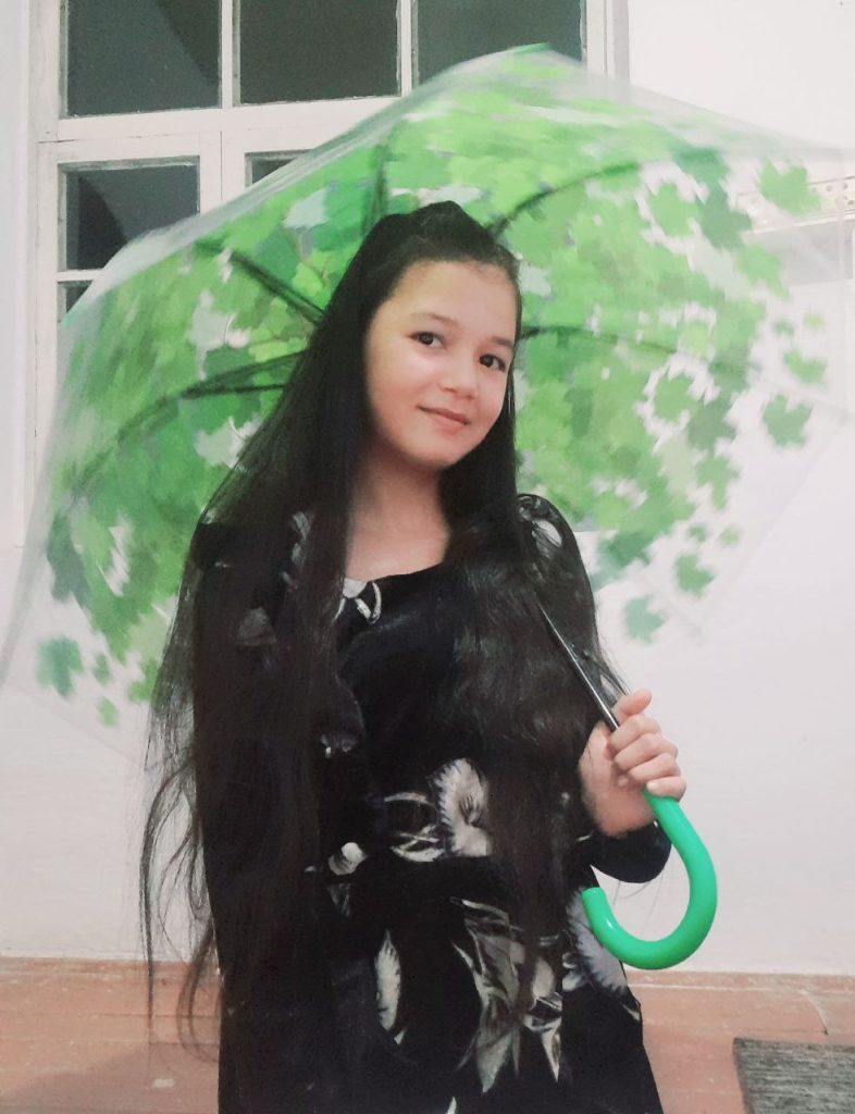 Central Asian teen girl with long dark hair and brown eyes in a dark black blouse holding a green and white umbrella. 