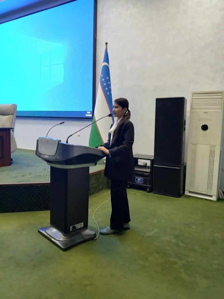 Central Asian woman with dark hair in a ponytail speaks at a podium in a room with desks and a projector. 