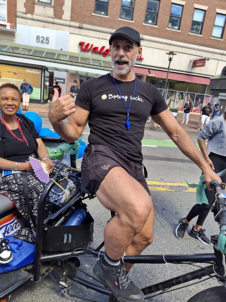 White man in a tee shirt that reads "Boring Sucks" and jeans and a black baseball cap gives a thumbs up to the camera. He's on a bike and has strong legs. 