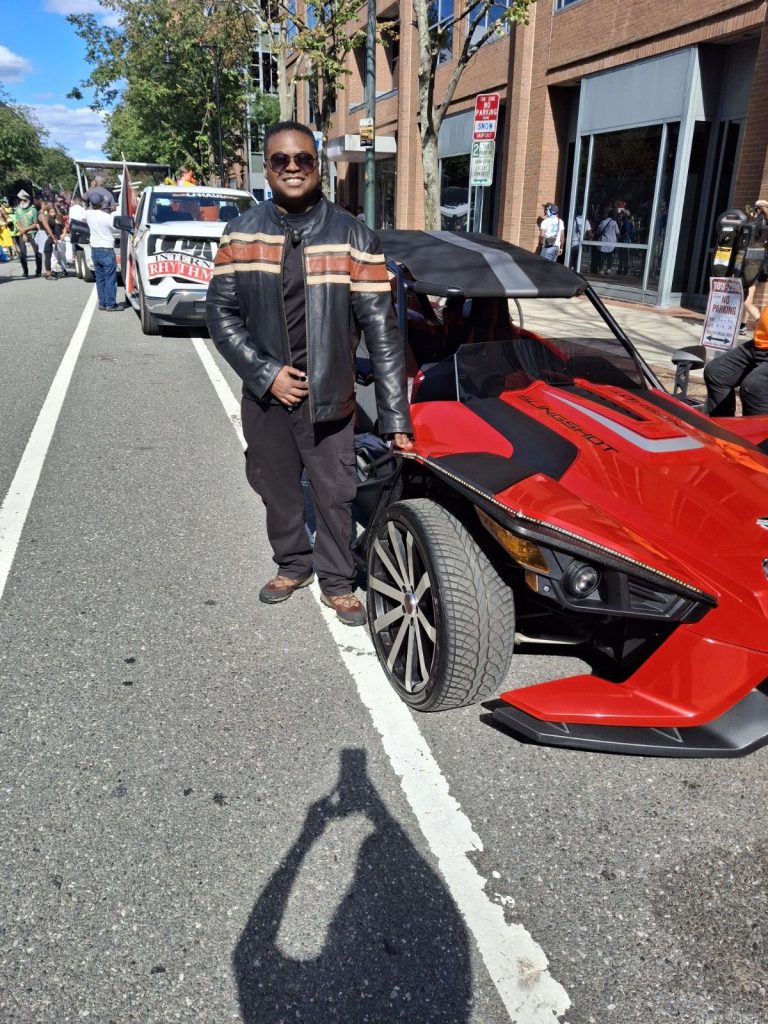 Black man in a jacket, black pants, sunglasses and sandals poses by a red sports car. 