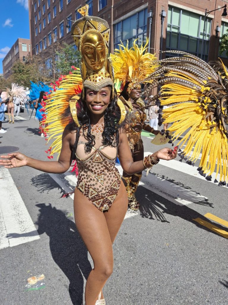 Young Black woman in a sequined costume with a yellow mask and headdress dances in a city street. 