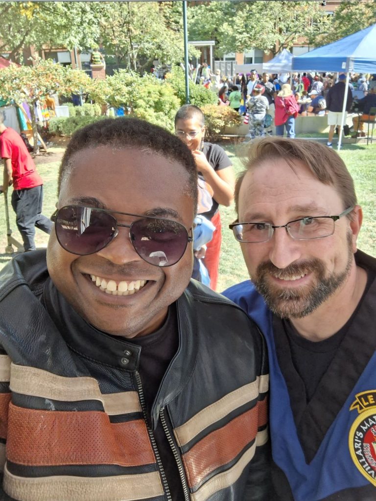 Two young middle aged men stand next to each other, one is white and the other black. They both have glasses on. Lots of other people and grass and trees are in the background. 