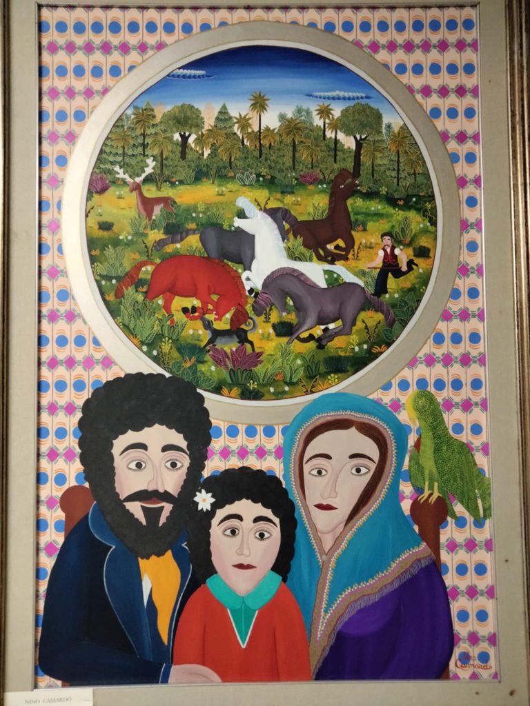 Flat image of a dark haired family, the man with a coat and a beard and sideburns and the woman with her hair covered and a jacket, and the child with a flower in her hair. Above them is a green parrot and a medallion window of a grassy meadow with trees, horses, a small figure of a man, and some other animals and trees and blue sky. 