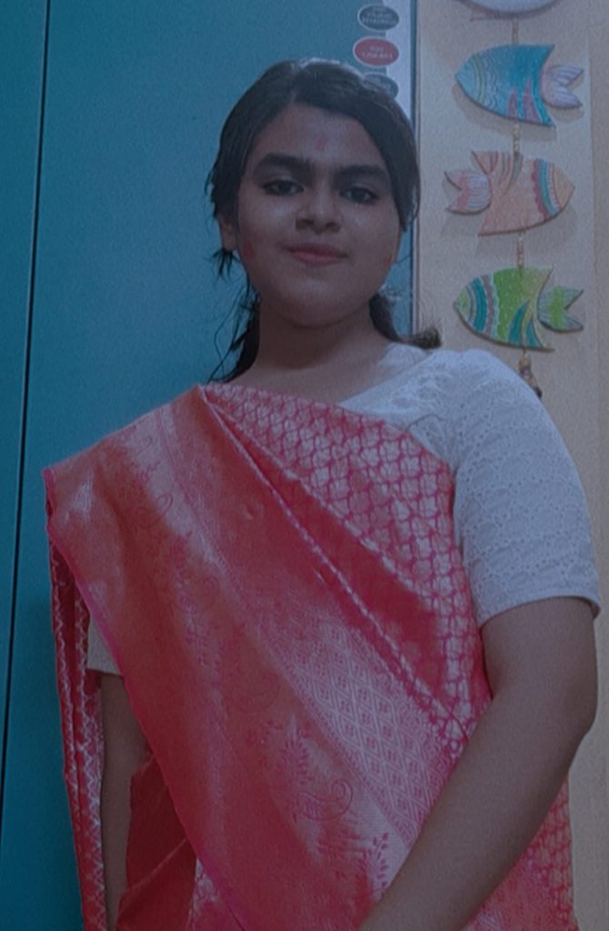 Young South Asian woman with short dark hair, brown eyes, and a patterned red and white sari. She's standing in front of a door with fish decals. 