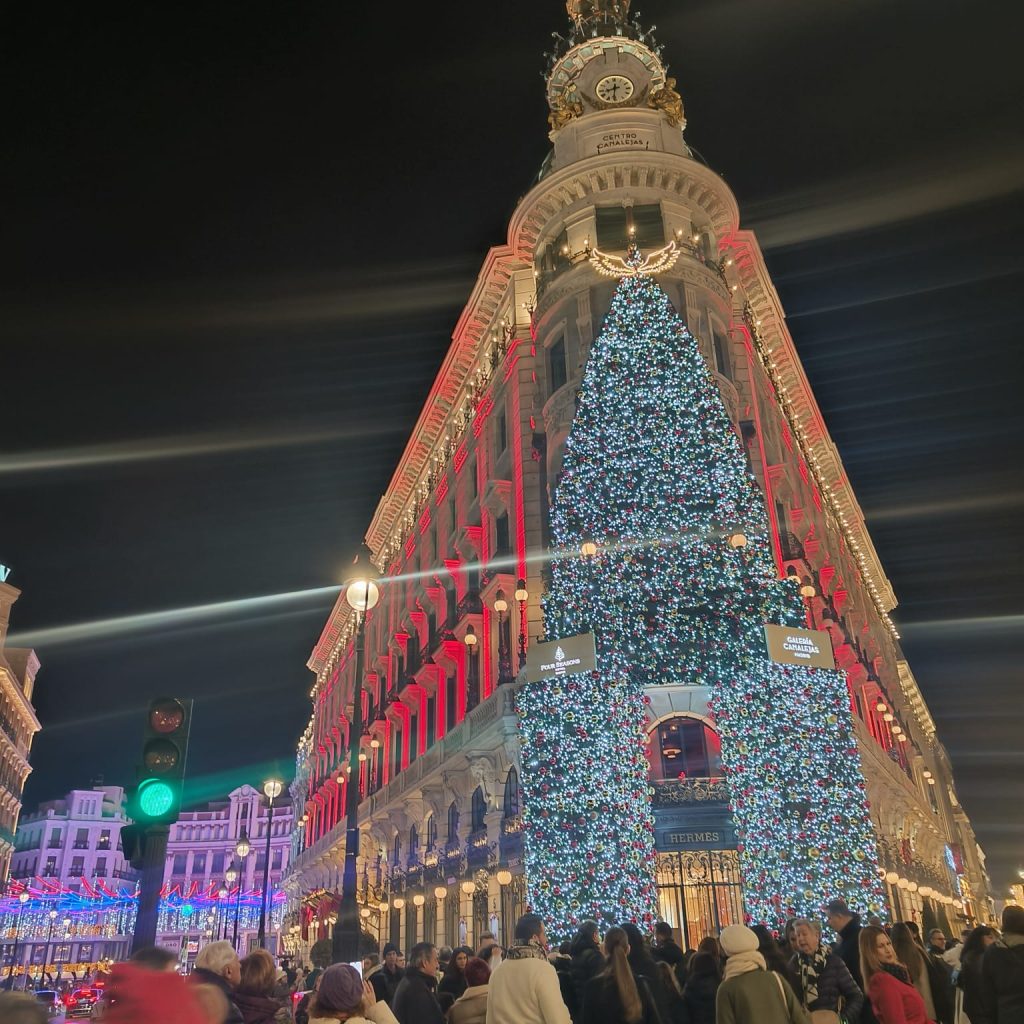 Large city building lit up like a Christmas tree at night for the holidays. People gathering around and a green streetlight.