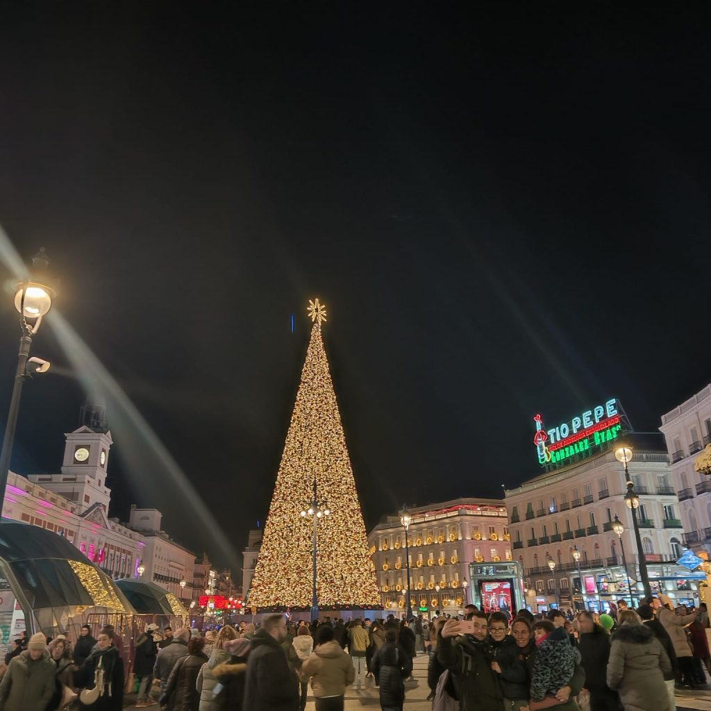 Large Christmas tree lit up with red balls and white lights and a star in the city at night. Lanterns, people, and buildings out.