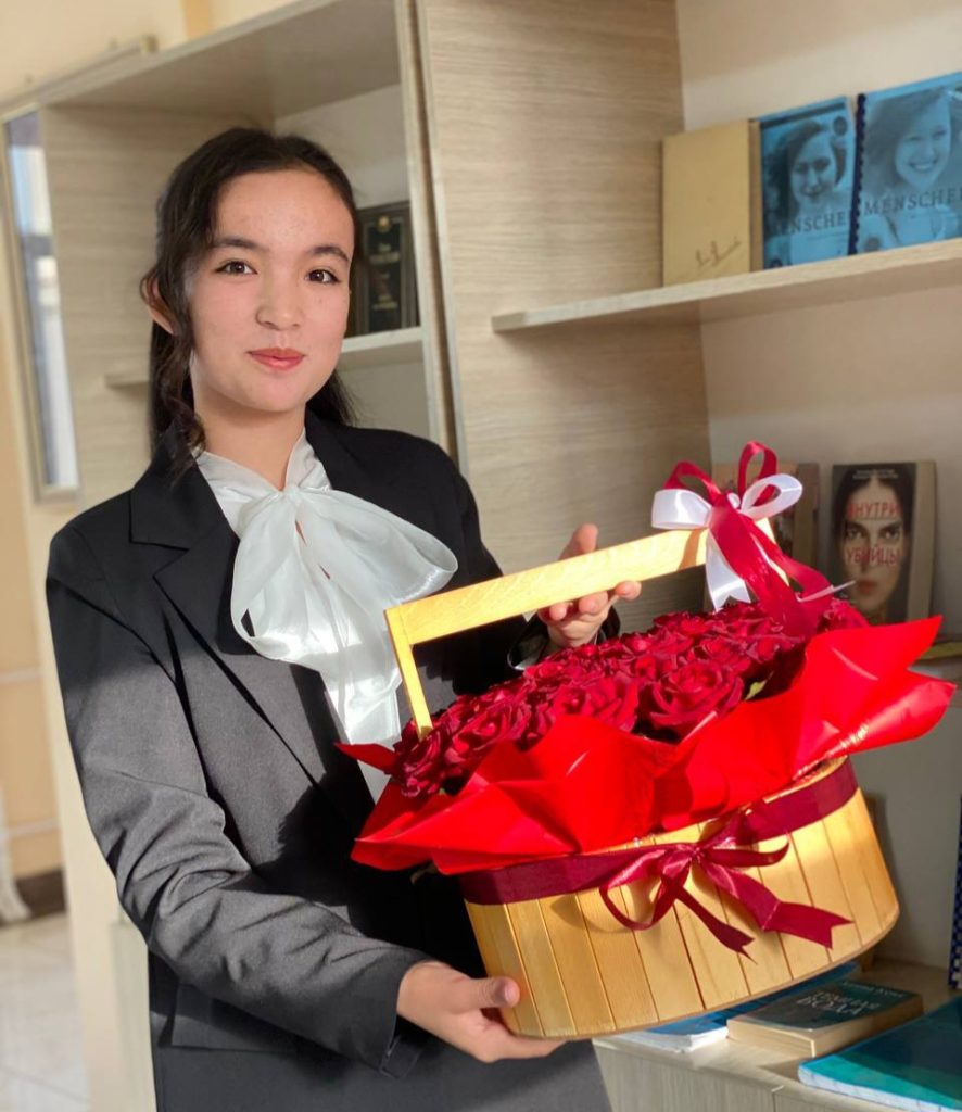 Central Asian young woman with long curly dark hair, black coat, and white ruffled blouse. She's holding a wooden basket with red roses and standing in front of a bookshelf filled with books. 