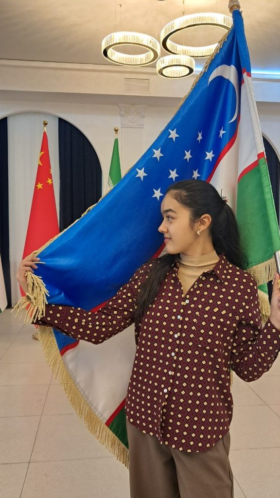 Central Asian teen girl with long dark hair in a barrette behind her head, brown eyes, earrings, and a burgundy and tan patterned top and tan pants standing in front of the Uzbek flag. 