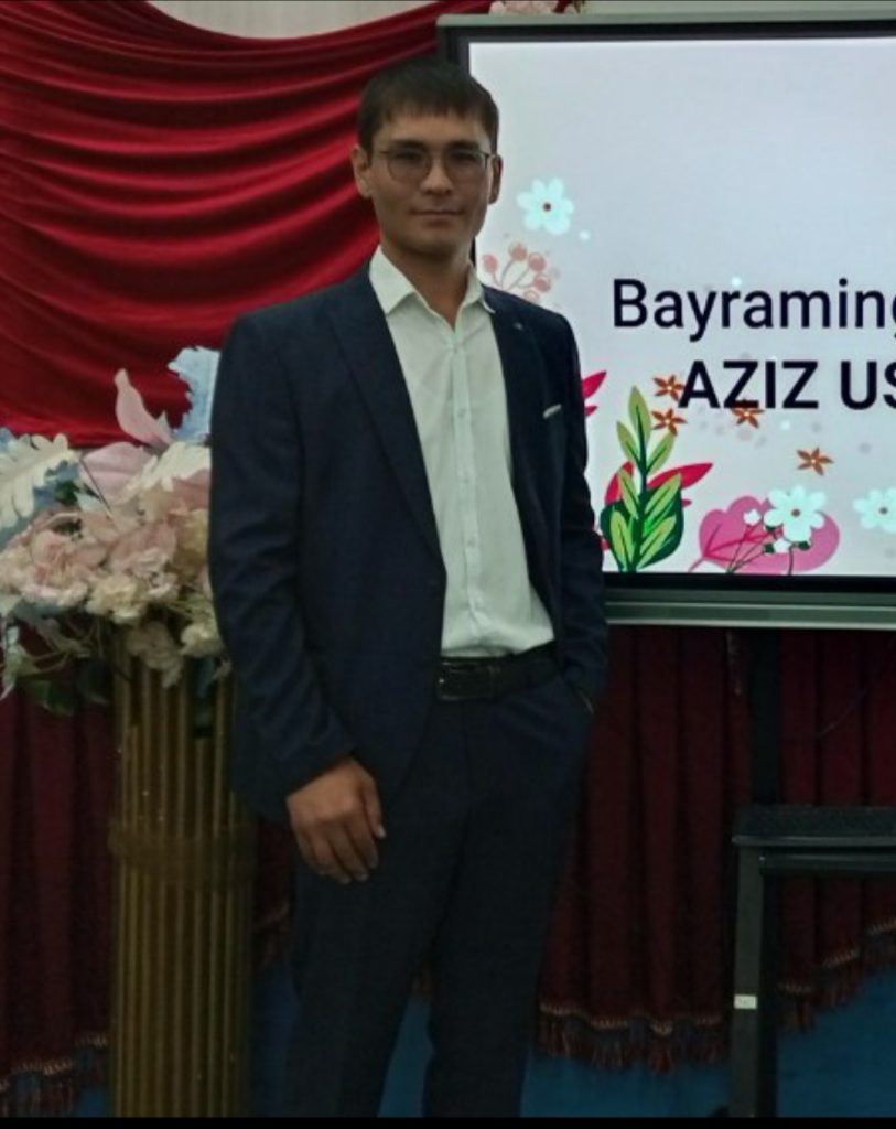 Young Central Asian man with short dark hair, reading glasses, clean shaven and a black coat and suit at a ceremony with a power point slide announcing him and red drapery and flowers. 