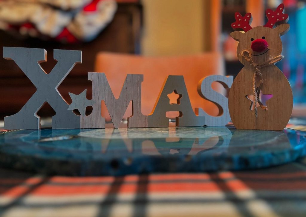 Brown wooden letters on a table spelling out "Xmas" with a wooden reindeer figure with a star on its belly and red antlers on the right of "Xmas." 