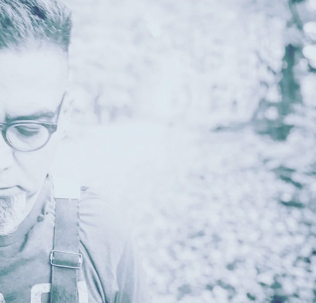 Image of a middle aged, light skinned man with reading glasses and overalls to the far left of a black and white photo. Unclear background. 