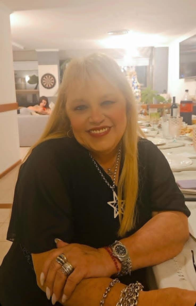 Light skinned, blonde/red haired Latina woman, smiling, with lipstick and earrings. She's got a necklace and a black v-neck top and is in front of a living room and a dining room with some food and drinks on the table and a woman sitting on the couch. She's wearing a watch and rings and bracelets and a star necklace. 